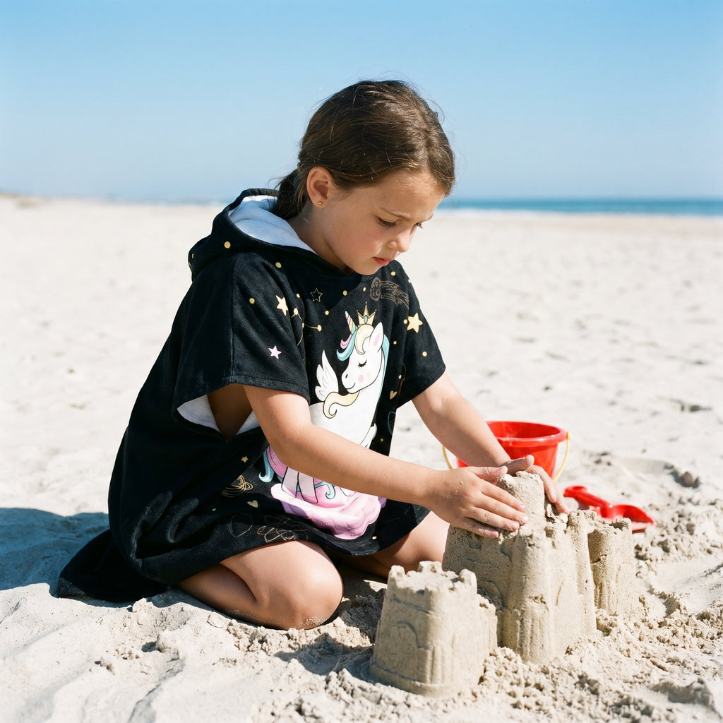 Poncho serviette a capuche motif licorne étoilée, porté par une enfant construisant un château de sable.