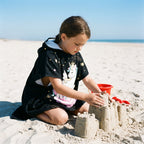 Poncho serviette a capuche motif licorne étoilée, porté par une enfant construisant un château de sable.