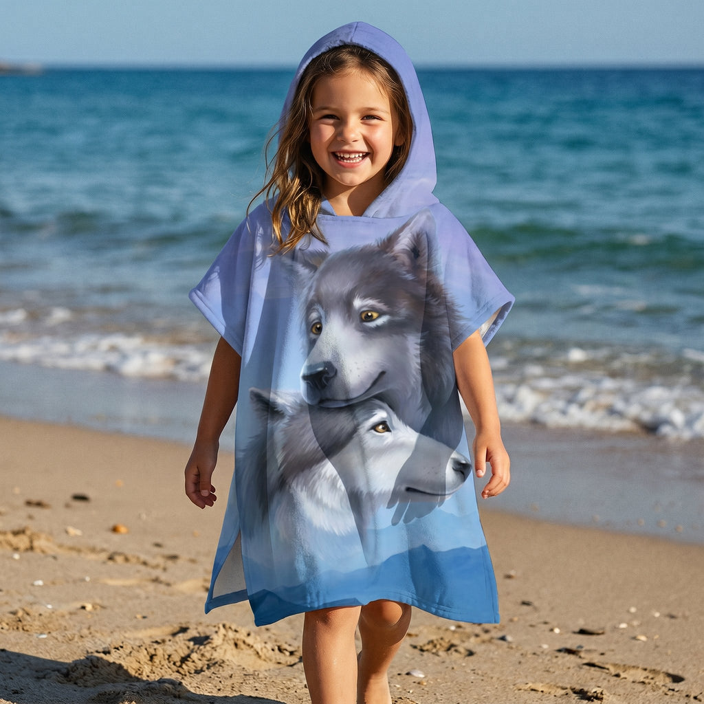 Poncho serviette a capuche motif loups, porté par une petite fille souriante marchant sur le sable fin de la plage.