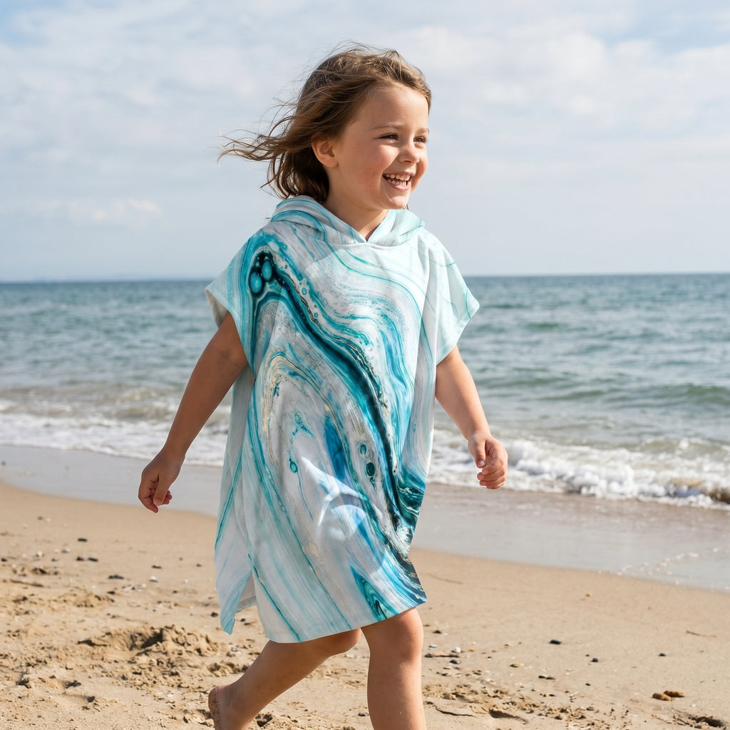 Poncho serviette a capuche motif marbre bleu ciel, porté par une enfant souriante marchant sur le sable de la plage.