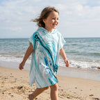 Poncho serviette a capuche motif marbre bleu ciel, porté par une enfant souriante marchant sur le sable de la plage.