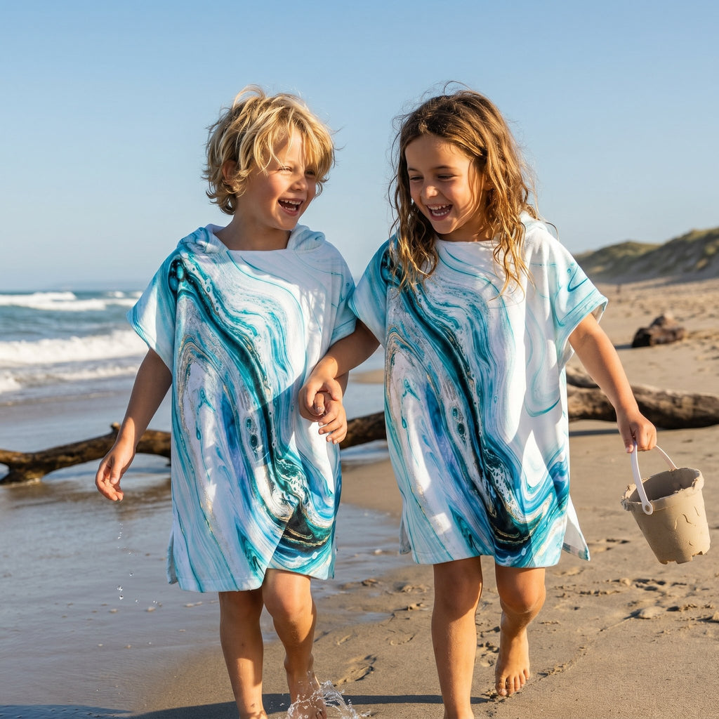 Poncho serviette a capuche motif marbre bleu ciel, porté par deux enfants rieurs se tenant la main sur le rivage.