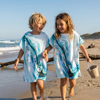 Poncho serviette a capuche motif marbre bleu ciel, porté par deux enfants rieurs se tenant la main sur le rivage.