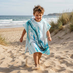 Poncho serviette a capuche motif marbre bleu ciel, porté par un enfant courant joyeusement sur le sable fin de la plage.