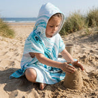 Poncho serviette a capuche motif marbre bleu ciel, porté par une enfant assise sur le sable, construisant un château.