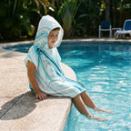 Poncho serviette a capuche motif marbre bleu ciel, porté par un enfant assis au bord d'une piscine, les pieds dans l'eau.