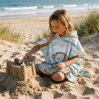 Poncho serviette a capuche motif marbre cuivré, porté par une enfant assise sur le sable.