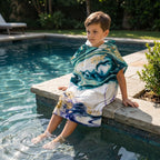 Poncho serviette a capuche motif marbre nuit dorée, porte par un garcon assis au bord d'une piscine.