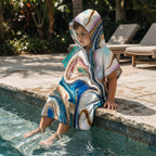 Poncho serviette a capuche motif marbre tourbillon cuivré, porté par un enfant assis au bord d'une piscine.