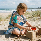 Poncho serviette a capuche motif marbre tourbillon cuivré, porté par une enfant assise sur le sable jouant.