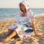 Poncho serviette a capuche motif Fonds marins colorés, porté par une femme assise de profil sur le sable.