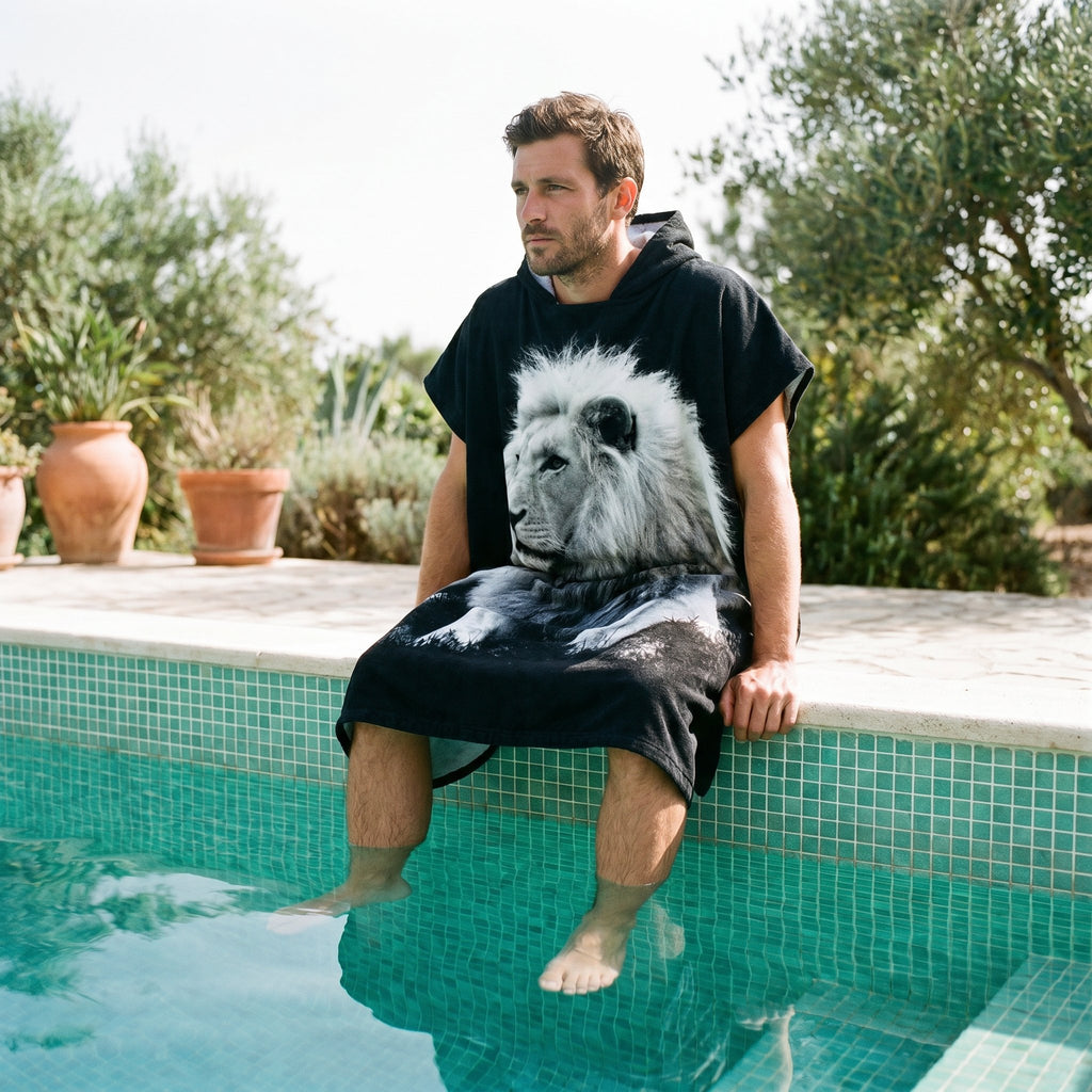 Poncho serviette à capuche motif lion noir et blanc, porté par un homme assis au bord d'une piscine.