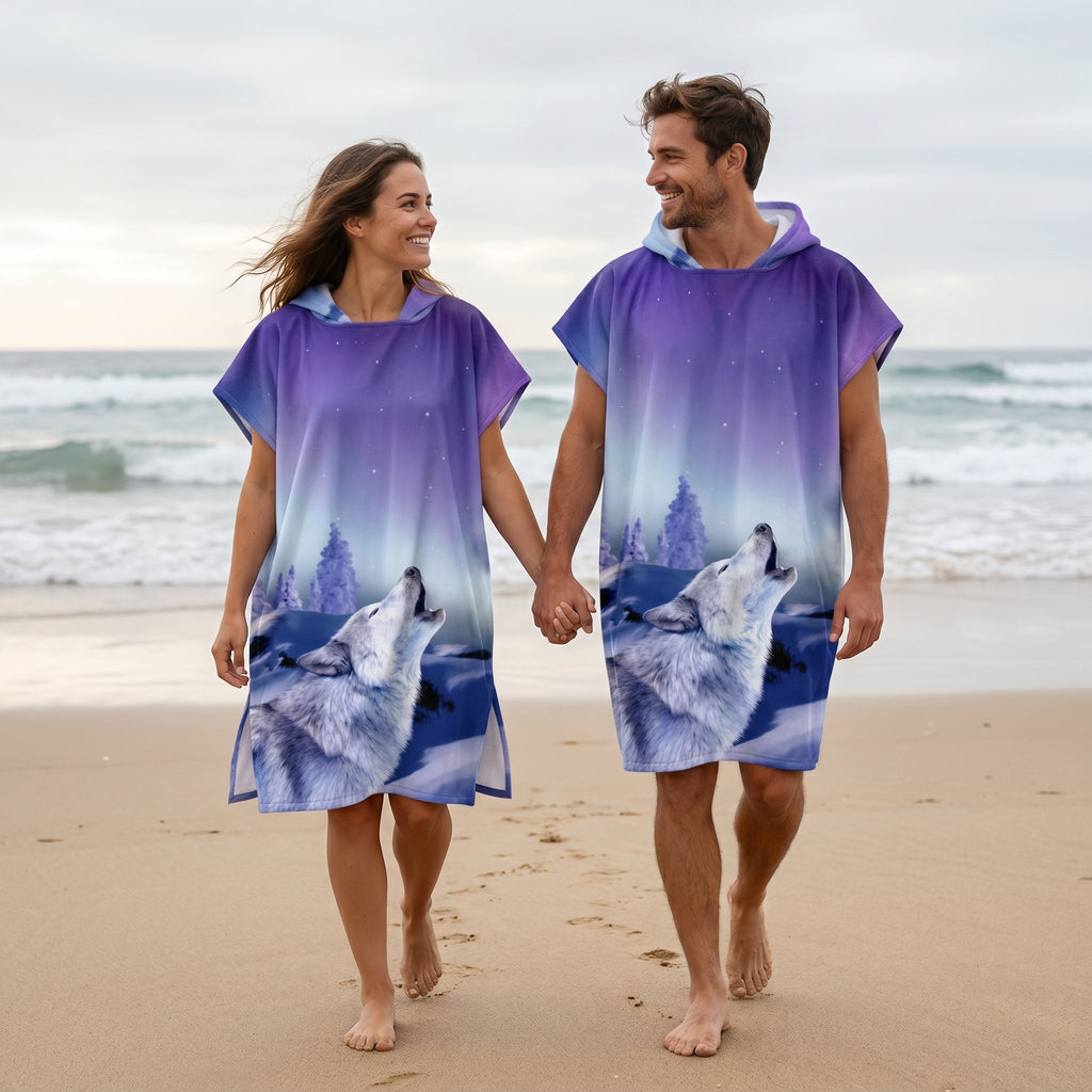 Poncho serviette a capuche motif loup aurore boreale, porte par un couple marchant main dans la main sur une plage.