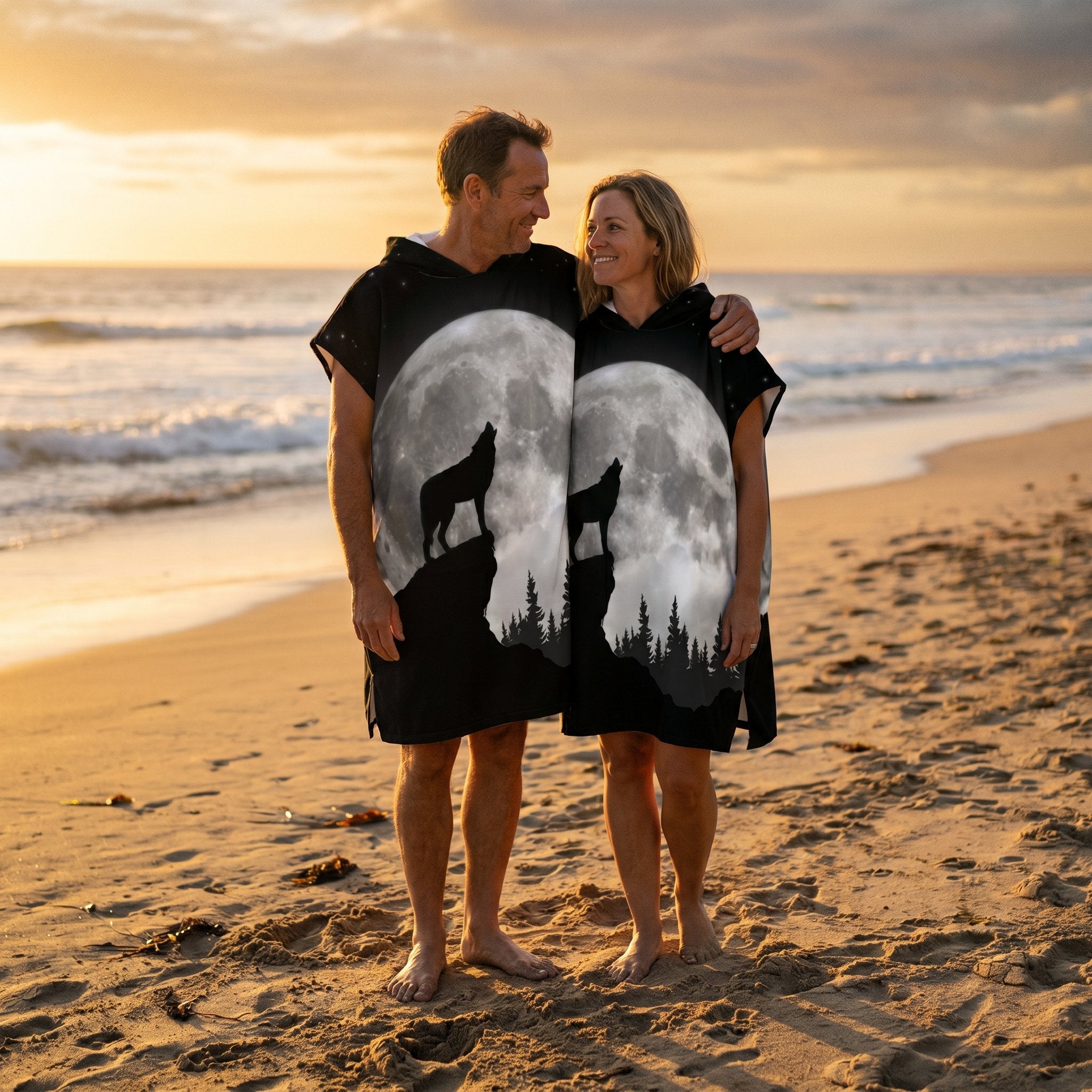 Poncho serviette à capuche motif loup et lune, porté par un couple souriant sur une plage au coucher du soleil, vue large.