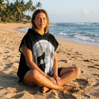 Poncho serviette à capuche motif loup et lune, porté par une femme assise en tailleur sur le sable d'une plage avec palmiers.