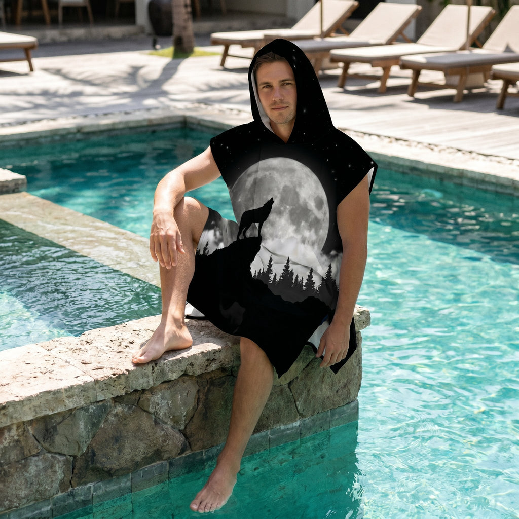 Poncho serviette à capuche motif loup et lune, porté par un homme assis au bord d'une piscine, les pieds dans l'eau.