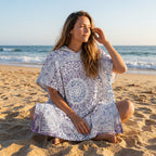 Poncho serviette à capuche motif mandala dentelle violet porté par une femme assise en tailleur sur le sable.