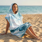 Poncho serviette a capuche motif marbre bleu givré, porté par une femme assise de profil sur le sable fin d'une plage.