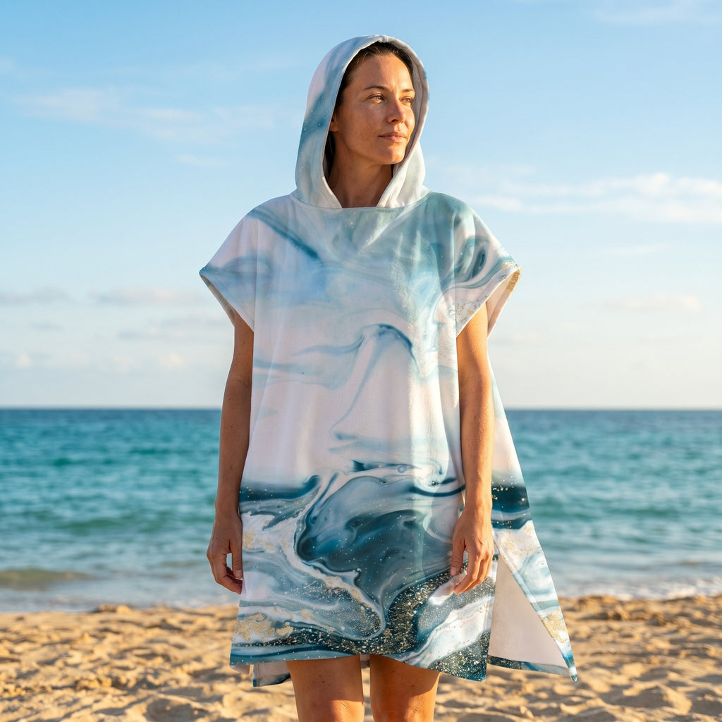 Poncho serviette a capuche motif marbre bleu givré, porté par une femme debout sur la plage, regardant vers la mer bleue.
