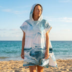 Poncho serviette a capuche motif marbre bleu givré, porté par une femme debout sur la plage, regardant vers la mer bleue.