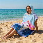 Poncho serviette a capuche motif marbre montagne rose, porte par une femme assise sur le sable fin.
