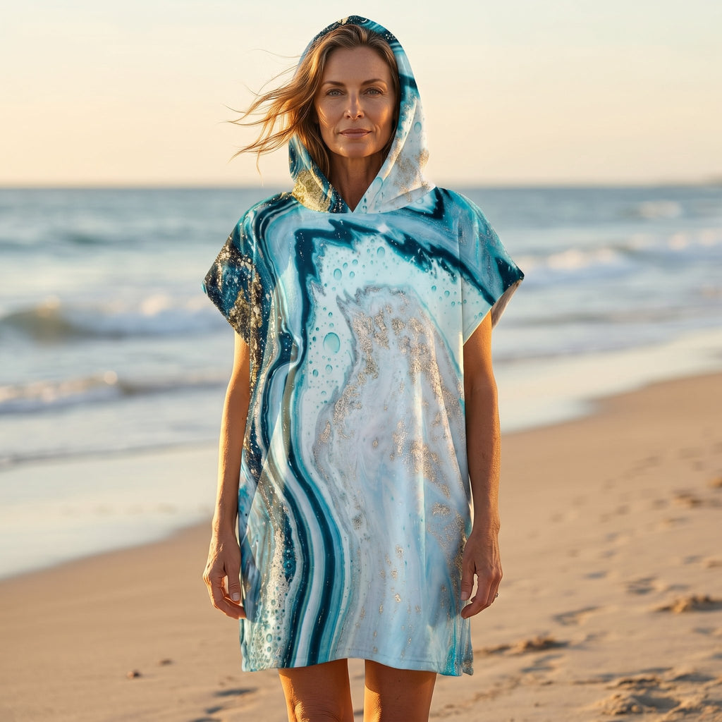 Poncho serviette à capuche motif marbre turquoise doré, porté par une femme debout sur le sable face à l'océan.