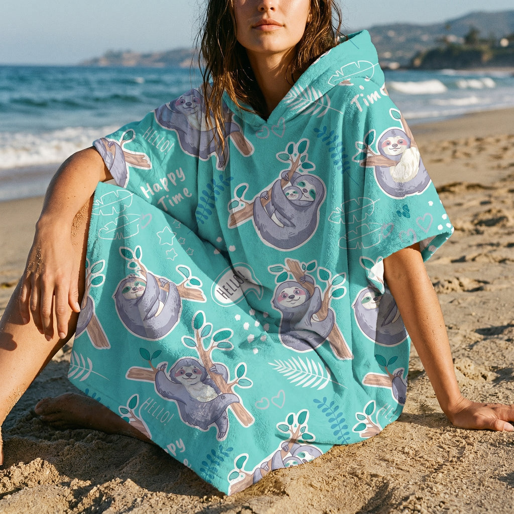 Poncho serviette a capuche motif paresseux turquoise, porte par une femme assise sur le sable pres de l'eau.