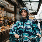 Poncho vélo à chevrons menthe, porté par une femme en gros plan, poussant son vélo devant une vitrine.
