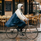 Poncho vélo crème et marine porté par une cycliste de profil, roulant devant une terrasse de café.