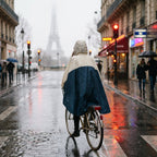 Poncho vélo crème et marine porté par une cycliste vue de dos, roulant sur une chaussée mouillée.