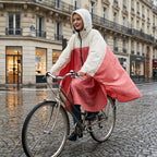 Poncho vélo crème et rose, porté par une femme souriante à vélo dans une rue pavée.