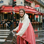 Poncho vélo crème et rose, porté par une femme debout à côté de son vélo devant un café.