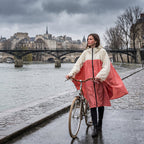 Poncho vélo crème et rose, porté par une femme marchant avec son vélo le long d'une rivière.