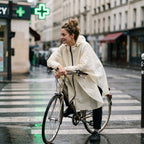 Poncho vélo crème uni, porté par une femme à vélo, arrêtée sur un passage piéton en ville.
