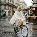 Poncho vélo crème uni, porté par une femme à vélo, roulant sur une rue pavée sous la pluie.