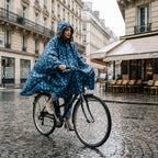 Poncho vélo motif fleurs bleu nuit, porté par une femme à vélo sur une rue mouillée, vue de côté.