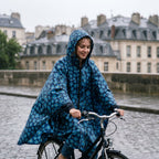 Poncho vélo motif fleurs bleu nuit, porté par une femme souriante à vélo, en plan rapproché sur un pont.