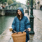 Poncho vélo motif fleurs bleu nuit, porté par une femme à vélo près d'un canal, tenant une tasse, vue de face.