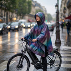 Poncho vélo irisé violet porté par une cycliste arrêtée à un passage piéton sur une avenue animée.