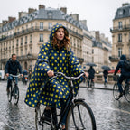 Poncho vélo motif pois moutarde porté par une femme rousse à vélo, vue de face sur une rue mouillée.