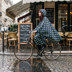 Poncho vélo motif pois moutarde porté par une cycliste rousse, vue de profil devant un café avec terrasse.