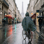 Poncho vélo motif pois moutarde porté par une femme à vélo, vue de dos sur une longue rue urbaine mouillée.