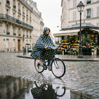 Poncho vélo motif pois moutarde porté par une femme à vélo, vue d'ensemble sur une rue pavée devant un fleuriste.